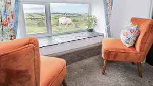 Une pièce avec deux fauteuils orange, une fenêtre donnant sur un champ où bêle un mouton blanc, et une plante en pot posée sur le rebord de la fenêtre.
