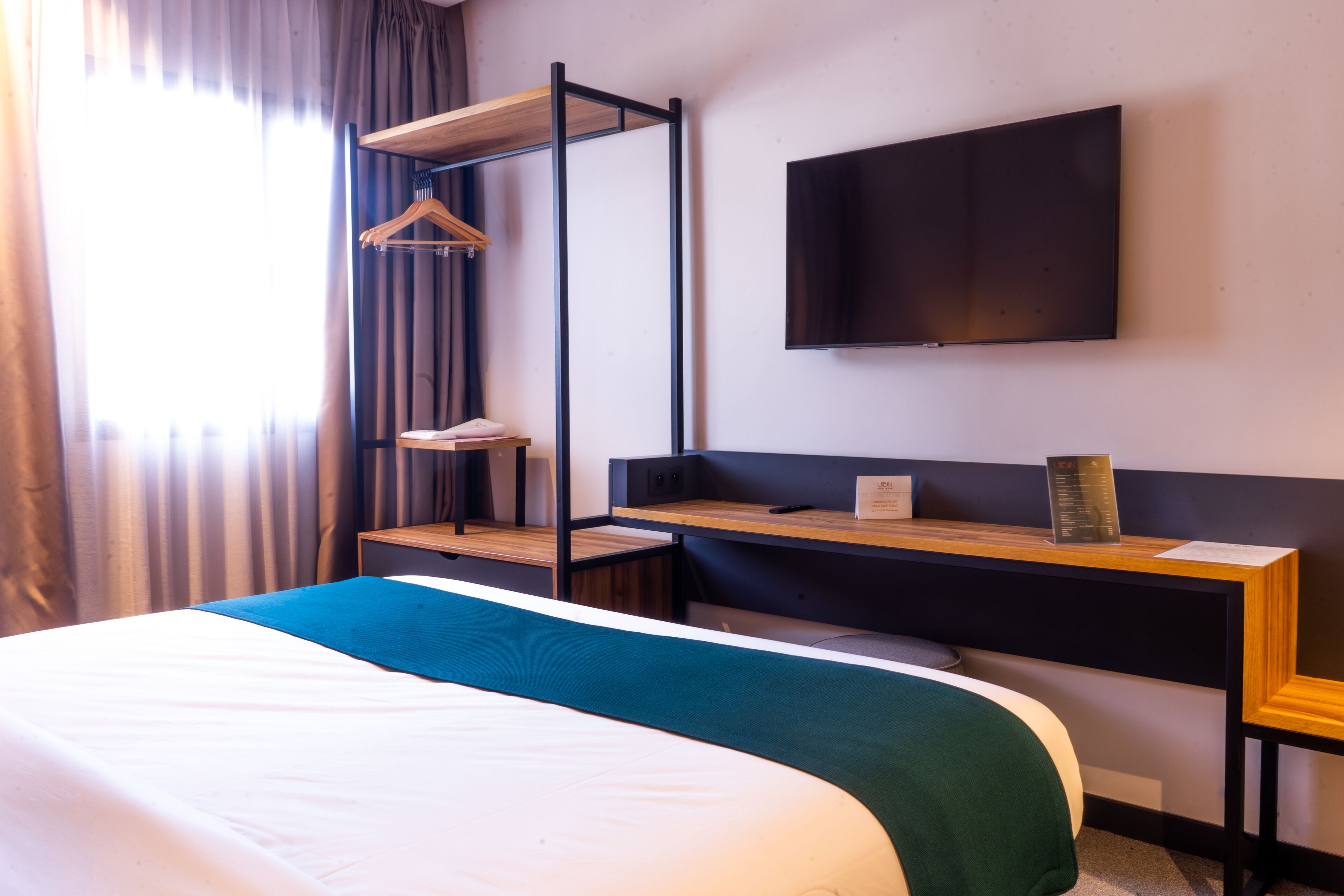 A hotel room with a bed, a desk, a TV mounted on the wall, and a window with curtains.