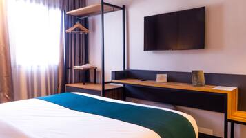 A hotel room with a bed, a desk, a TV mounted on the wall, and a window with curtains.