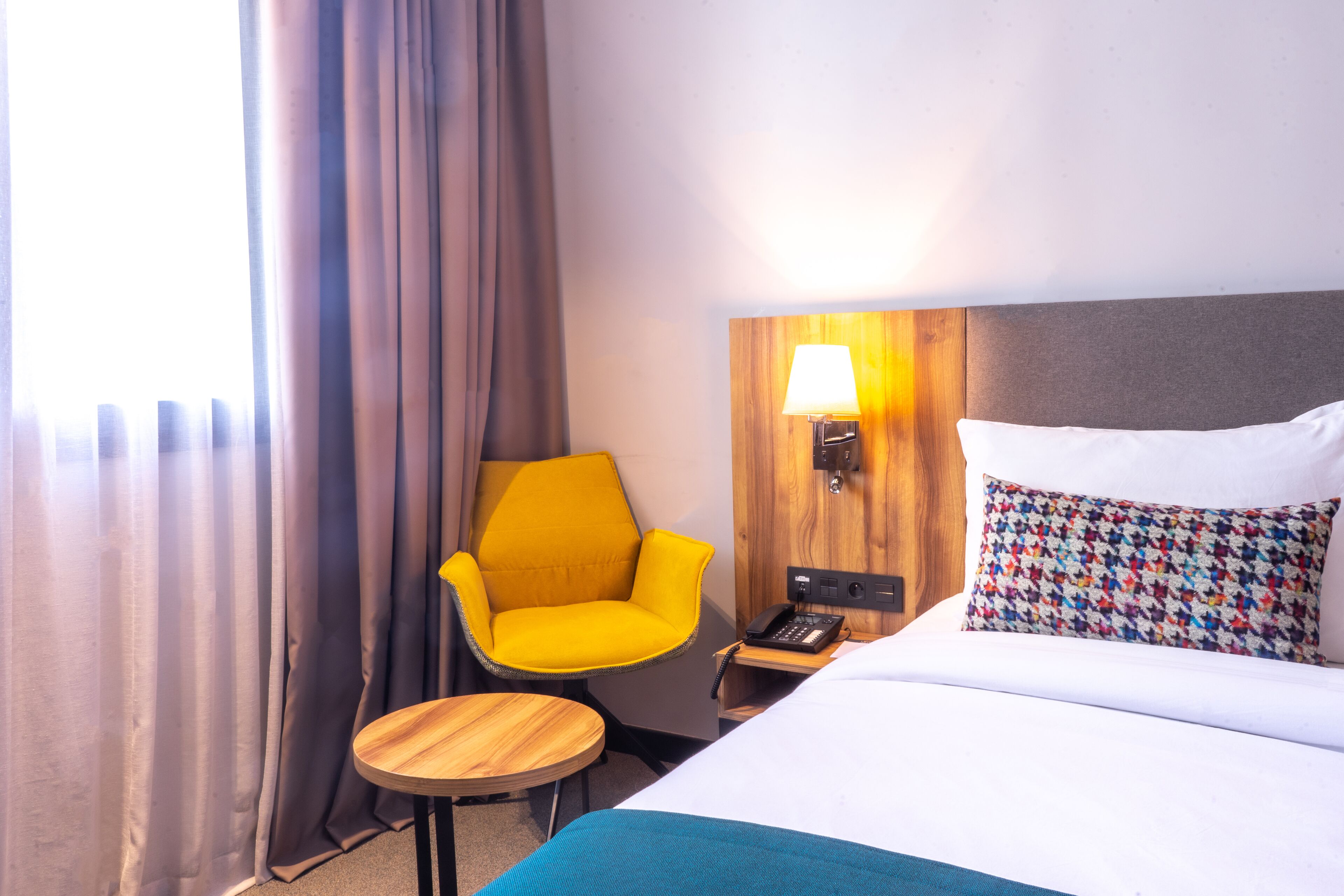 A hotel room with a bed, a nightstand, a lamp, a chair, and a small table.