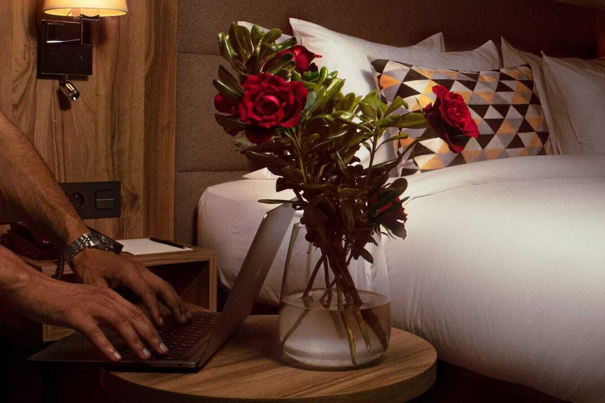 A hotel room with a bed, a laptop on a bedside table, and a vase with flowers.