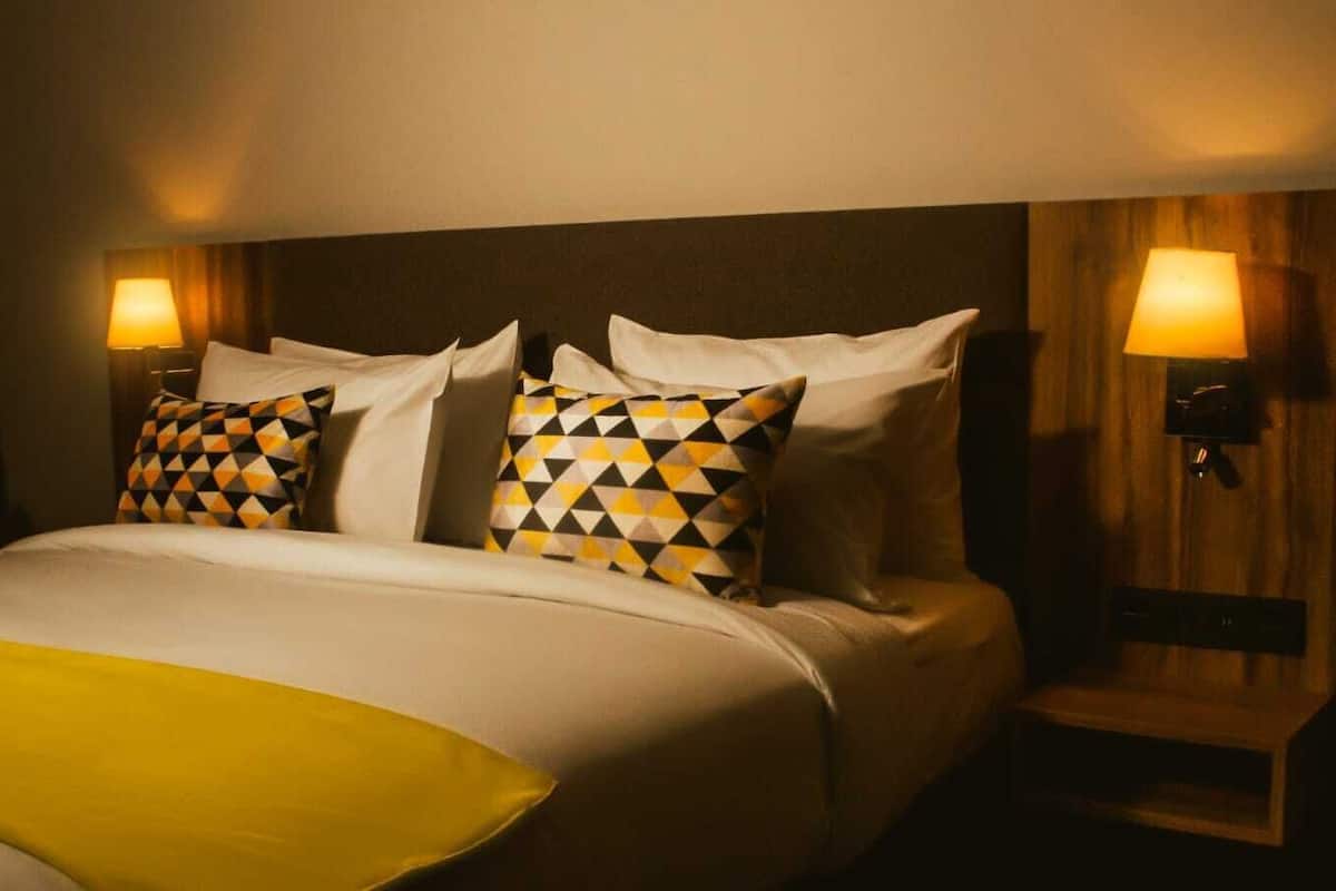 A hotel room with a bed, two pillows, a bedside table, and a lamp.