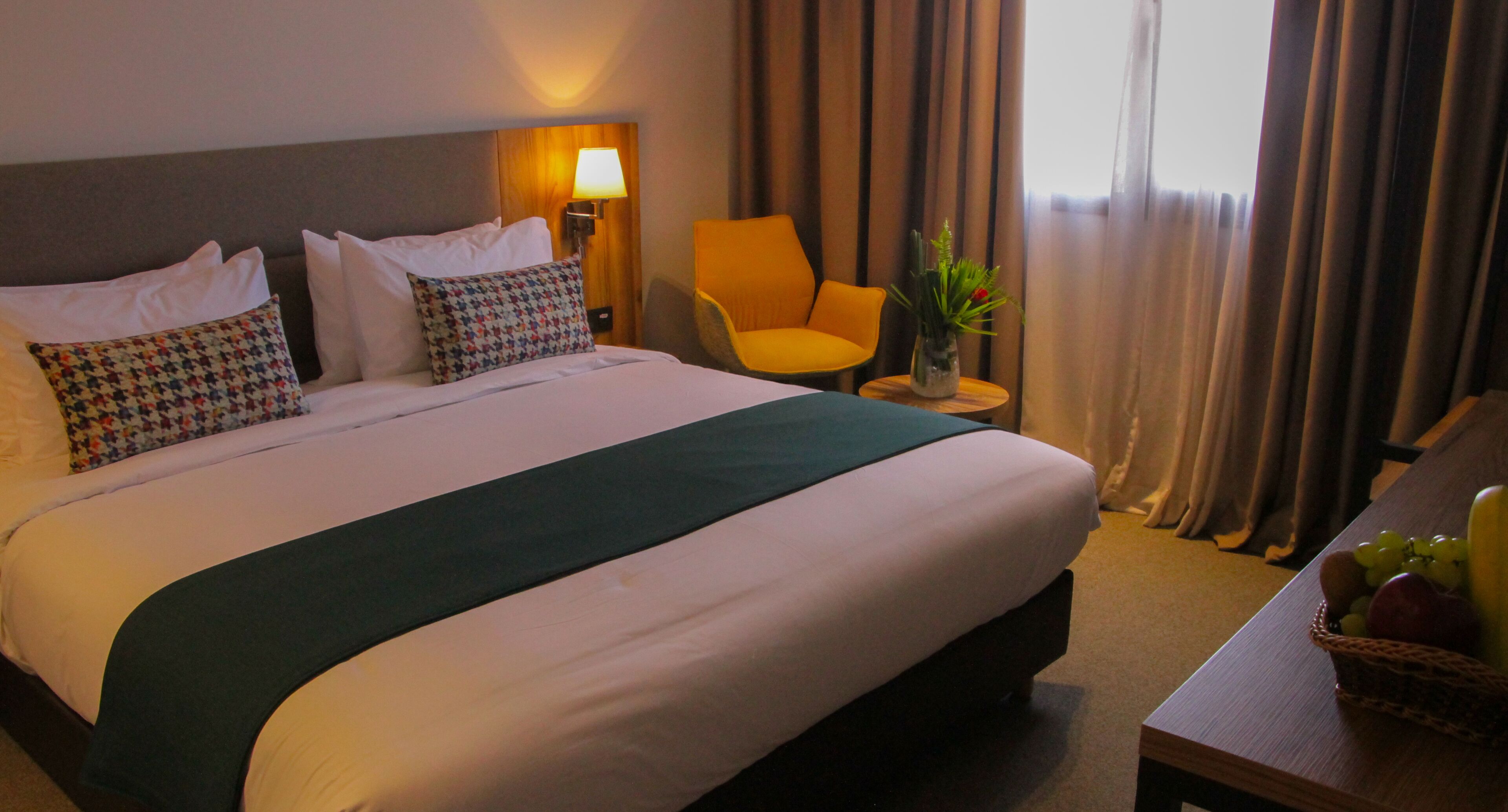 A hotel room with a large bed, a chair, a small table with a vase, and a fruit basket.