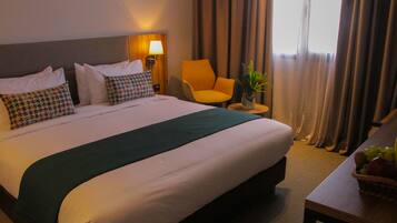 A hotel room with a large bed, a chair, a small table with a vase, and a fruit basket.