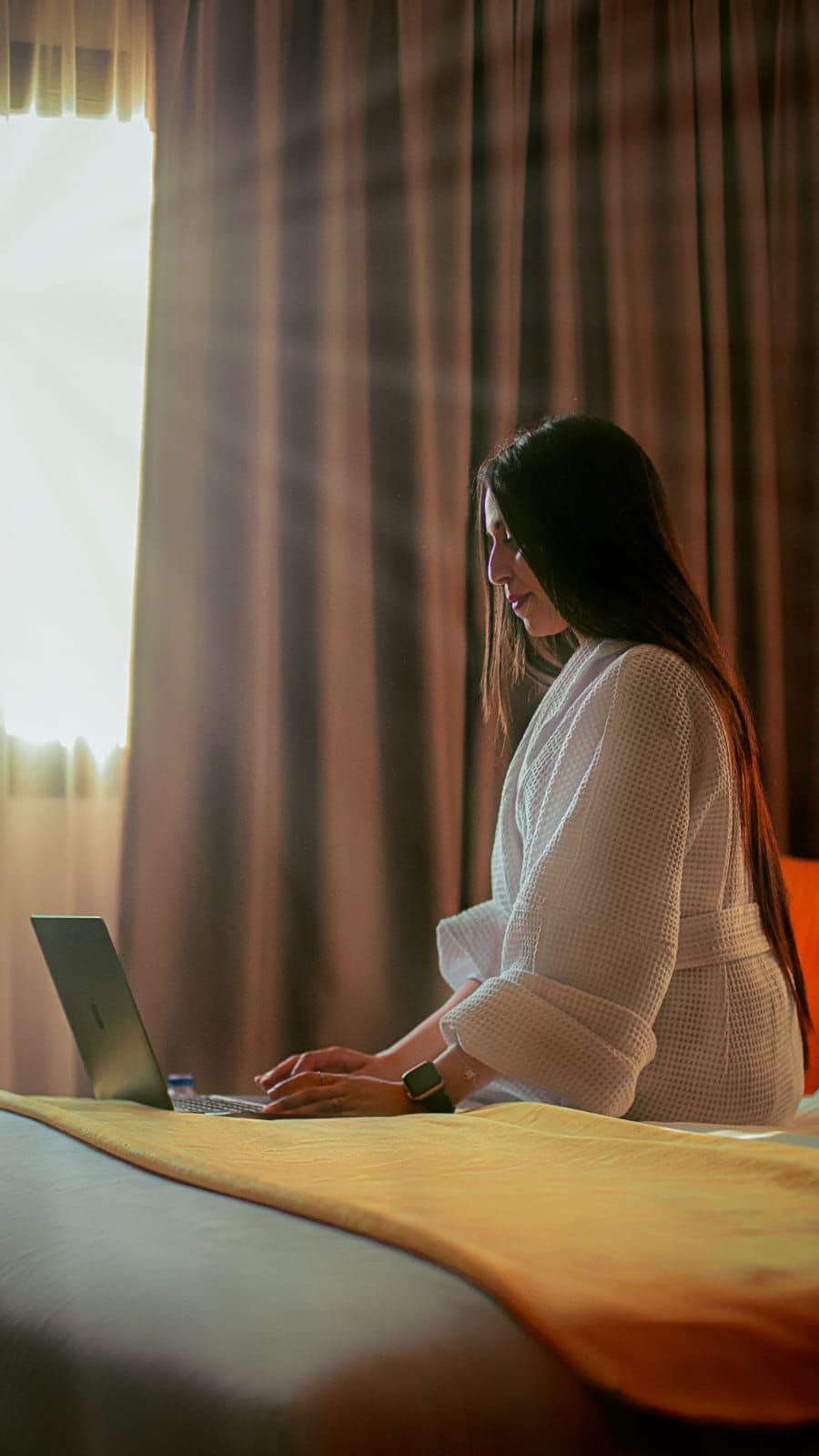 Una persona sentada en una cama con una computadora portátil, cortinas y una ventana al fondo.