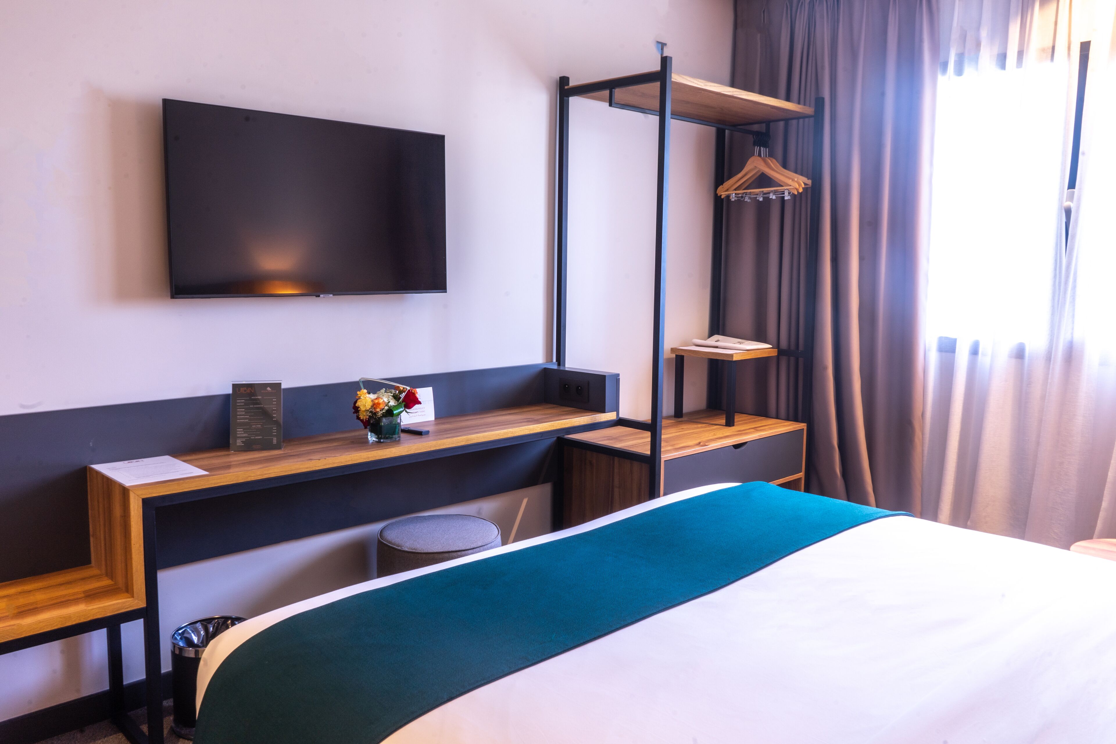 A hotel room with a bed, a TV mounted on the wall, a desk with a computer, and a hanging coat rack.