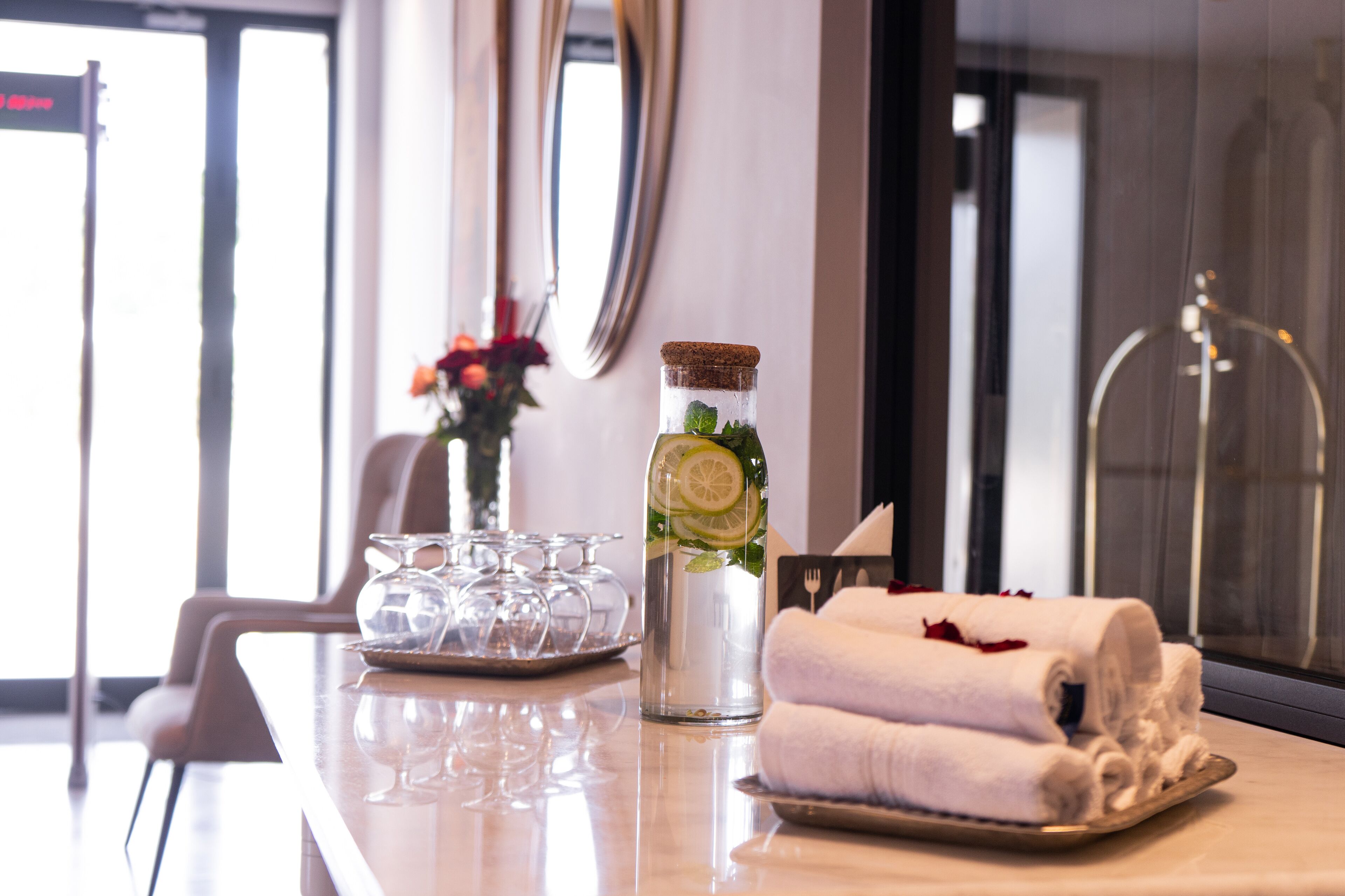 A hotel room with a glass table, a bottle of water with a lemon slice, a vase with flowers, and rolled towels.