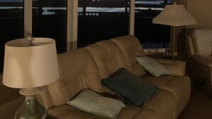 Un salon avec une grande fenêtre, un canapé, une lampe et une vue sur un coucher de soleil se reflétant sur une étendue d’eau.