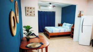 A small, compact room with a bed, a table with a plant, a refrigerator, and a ceiling fan.