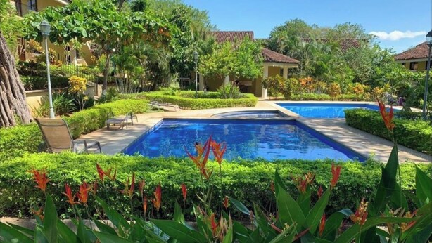 A pool area with a clear blue pool, surrounded by well-maintained greenery and a few lounge chairs.