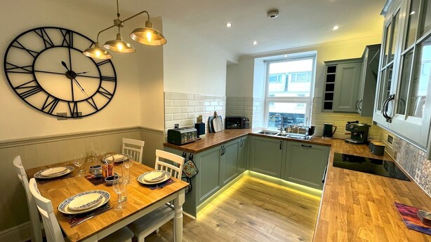 A modern kitchen with a dining area, featuring a large wall clock, wooden flooring, and a window providing natural light.