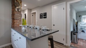 Une cuisine dotée d’un comptoir en granit, de meubles blancs et d’un mur d’accent en briques. On y voit deux panneaux encadrés accrochés au mur et une petite poubelle placée à droite.