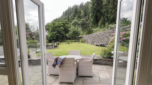 An outdoor patio with wicker furniture and a stone pathway leading to a grassy area with a stone wall and trees. - Pengwern Bach (Llanrwst)