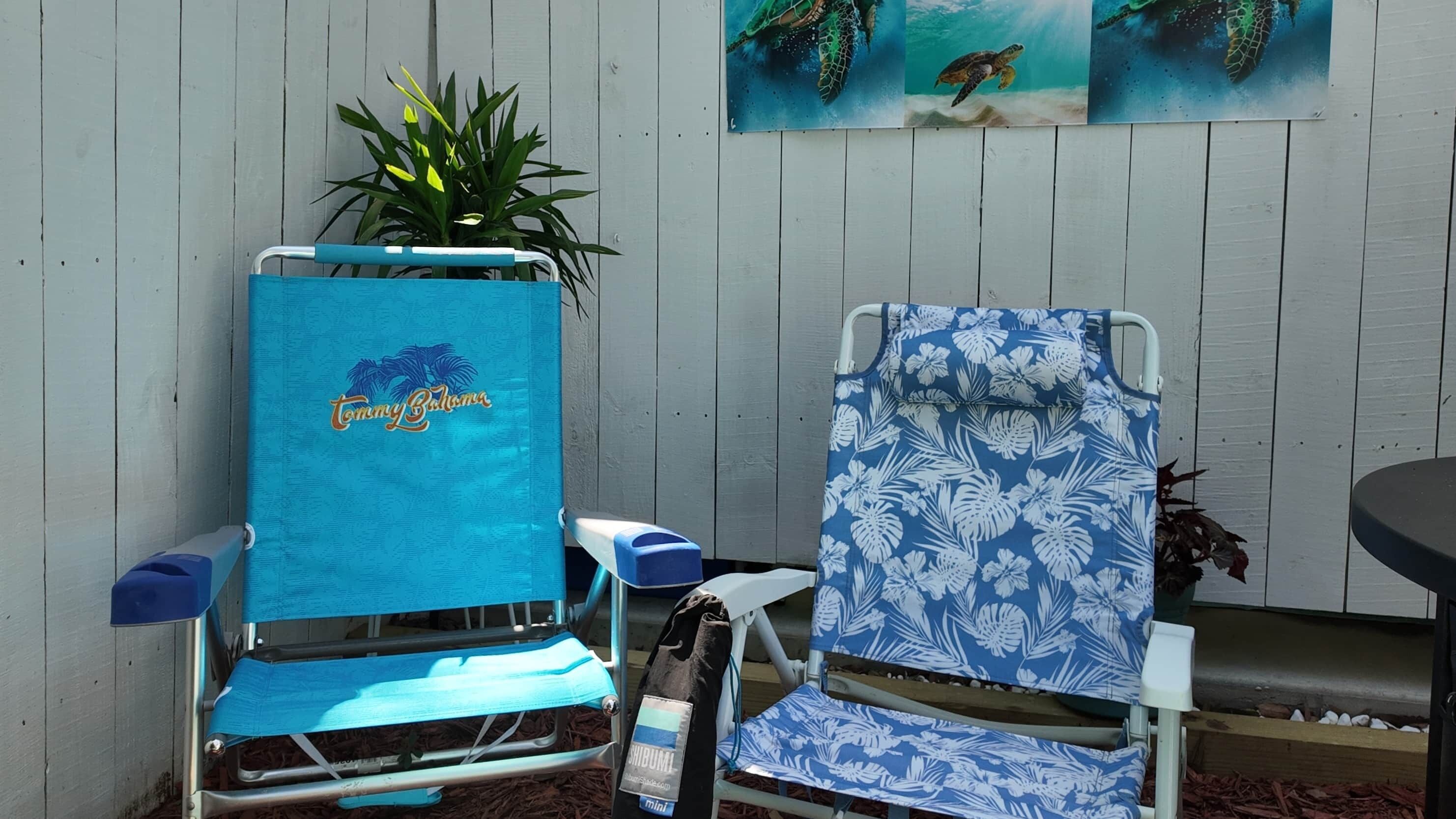 Zwei Strandstühle mit blauen Bezügen, einer mit dem Schriftzug 'Tommy Bahama' und der andere mit einem tropischen Blattmuster, vor einer Holzwand mit einem Blumentopf und einem Gemälde von Meeresschildkröten.