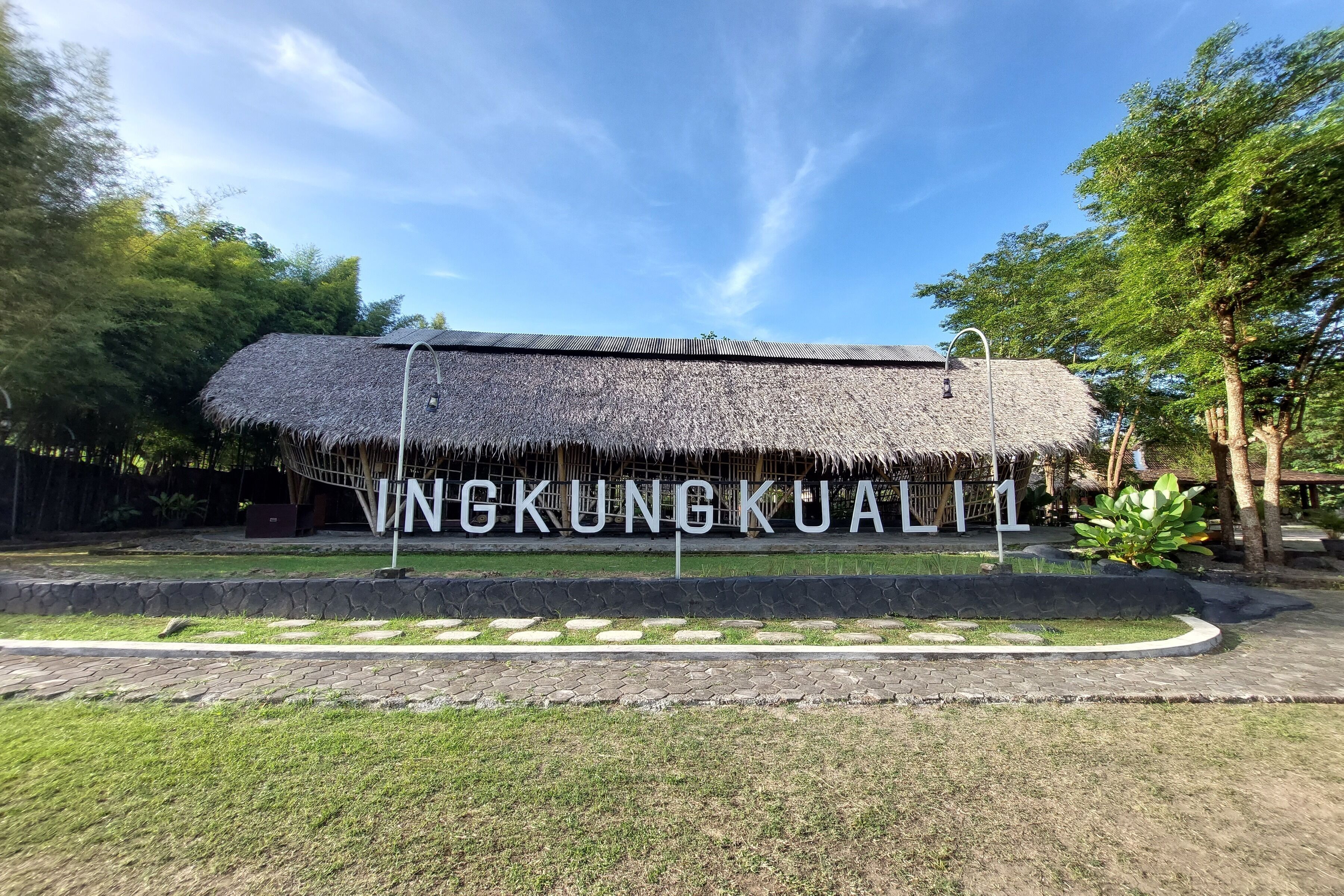 Uma estrutura com telhado de palha e uma placa com a inscrição "VNGKUNG KUA" em frente a uma área ajardinada com um caminho de pedra e gramado.