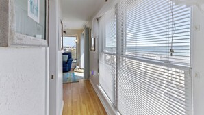 A hallway with white walls, wooden flooring, and large windows with blinds. There is a door at the end leading to a room with a blue chair and a small table. - Luxury, Ocean Front, Cargo Lift, 3rd Floor Penthouse KD0003 (Kill Devil Hills)