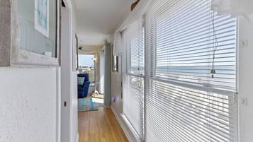 A hallway with white walls, wooden flooring, and large windows with blinds. There is a door at the end leading to a room with a blue chair and a small table.