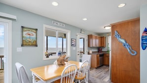 A dining area with a wooden table and white chairs, a kitchen with wooden cabinets, and a view of the ocean through a window.