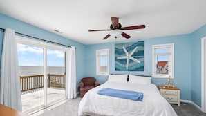 A bedroom with a large bed, a ceiling fan, a view of the ocean through a sliding door, and a starfish artwork on the wall.