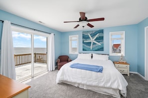 Quarto com cama grande, ventilador de teto, vista do oceano através de uma porta deslizante e uma obra de arte com estrela-do-mar na parede.