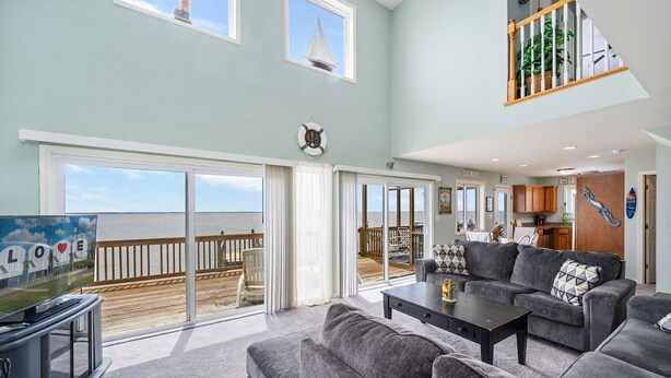 A spacious living room with a large sliding glass door leading to a deck with a view of the ocean. There is a gray sofa set and a small table with a decorative item.