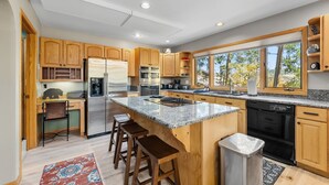 Una cocina moderna con armarios de madera, un refrigerador de acero inoxidable, una isla grande con taburetes de bar y vistas a árboles a través de las ventanas.