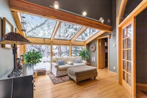 Un salon spacieux avec un plafond haut, des poutres en bois, une grande verrière, un ensemble de canapés, une table basse et une plante en pot.
