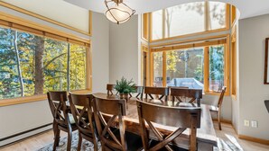 Un comedor con mesa y sillas de madera, ventanas amplias con vistas a árboles y una lámpara en el techo.