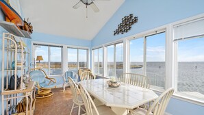 Habitación costera con una mesa grande y sillas, un ventilador de techo y vistas al mar a través de amplios ventanales.