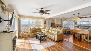 Un salón espacioso con un sofá seccional beige grande, una mesa de centro de madera y un ventilador de techo. Se ve una cocina con encimera de madera y taburetes de bar al fondo.