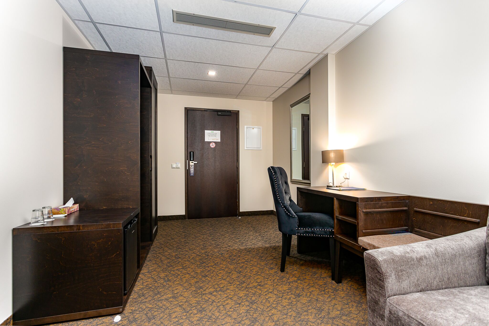 Habitación de hotel con un escritorio de madera oscura, un sillón con reposabrazos, un pequeño sofá, una lámpara de escritorio y una puerta con un cartel.