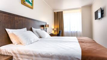 Een hotelkamer met een groot bed, een houten hoofdeinde, een bureau met een lamp, een televisie en een raam met gordijnen.