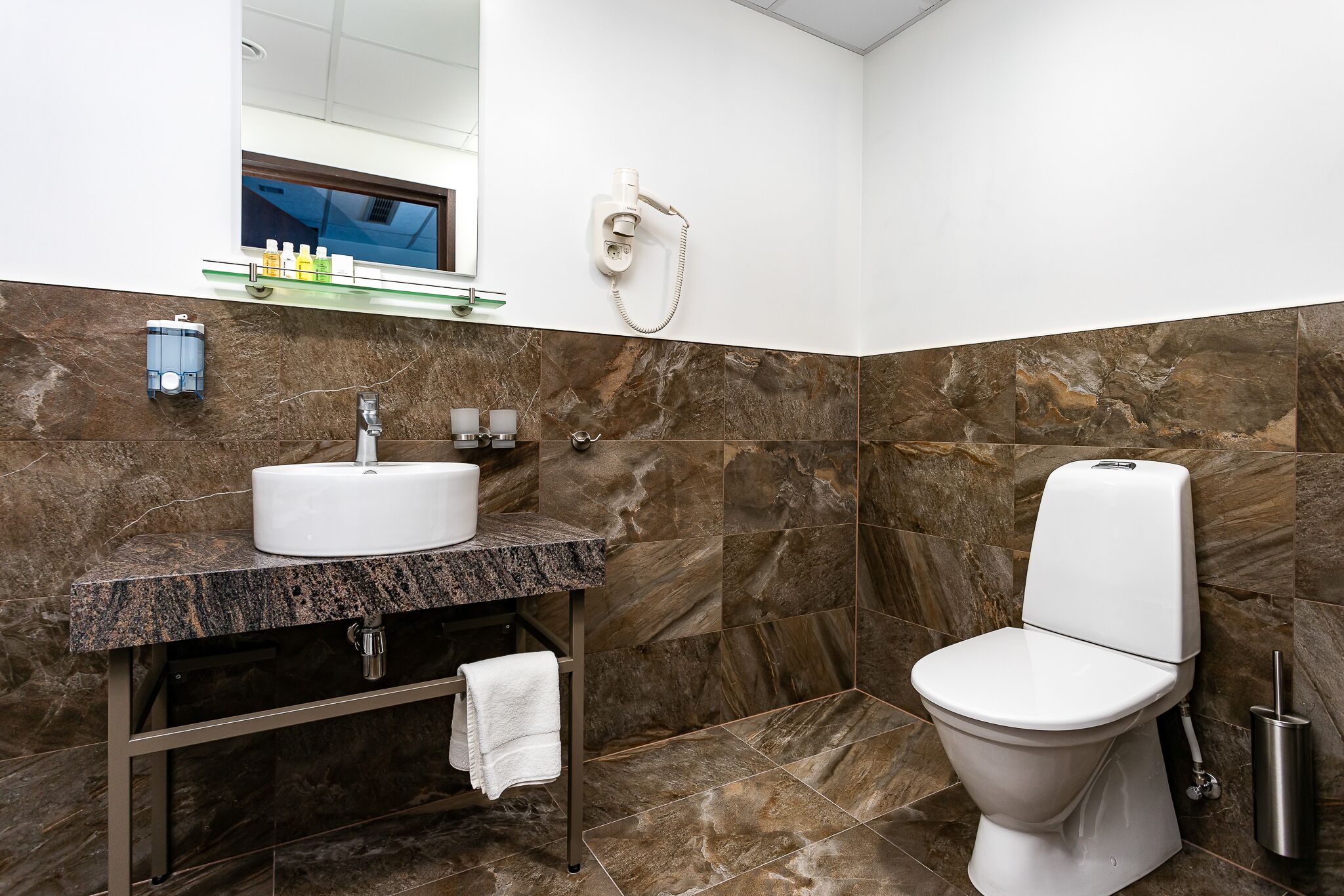 Une salle de bain avec un lavabo en marbre, des toilettes, une douchette à main et une étagère en verre où sont rangés les articles de toilette.