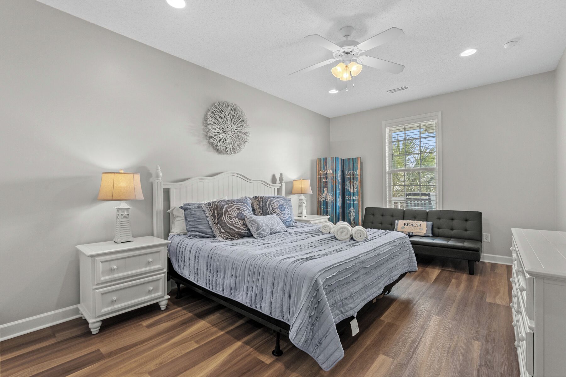 Une chambre à coucher comprenant un lit, des tables de chevet, une commode, un ventilateur de plafond et une fenêtre avec des stores.