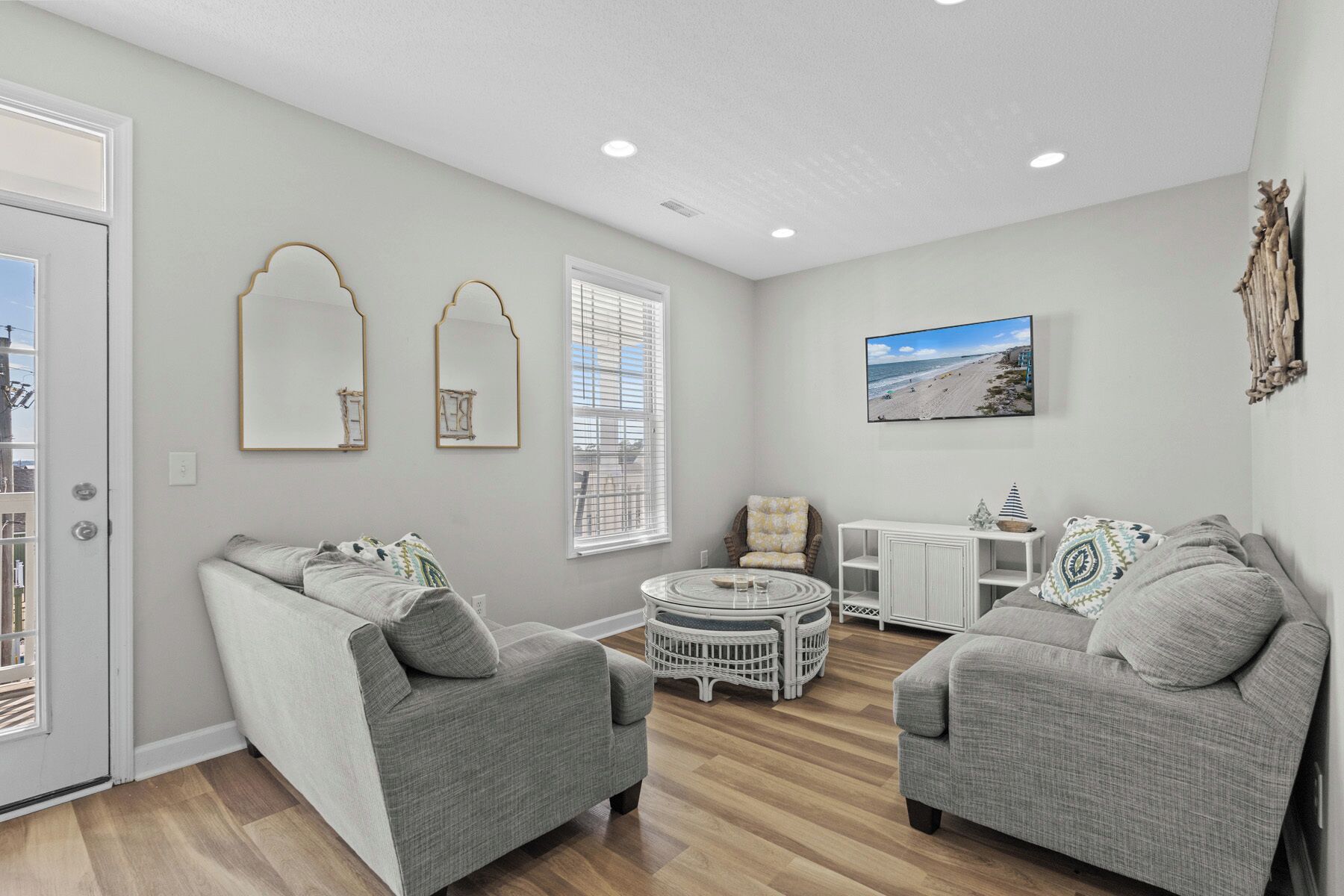 A modern living room with two grey sofas, a round coffee table, and a white side table. There are two wall-mounted mirrors and a beach-themed painting.