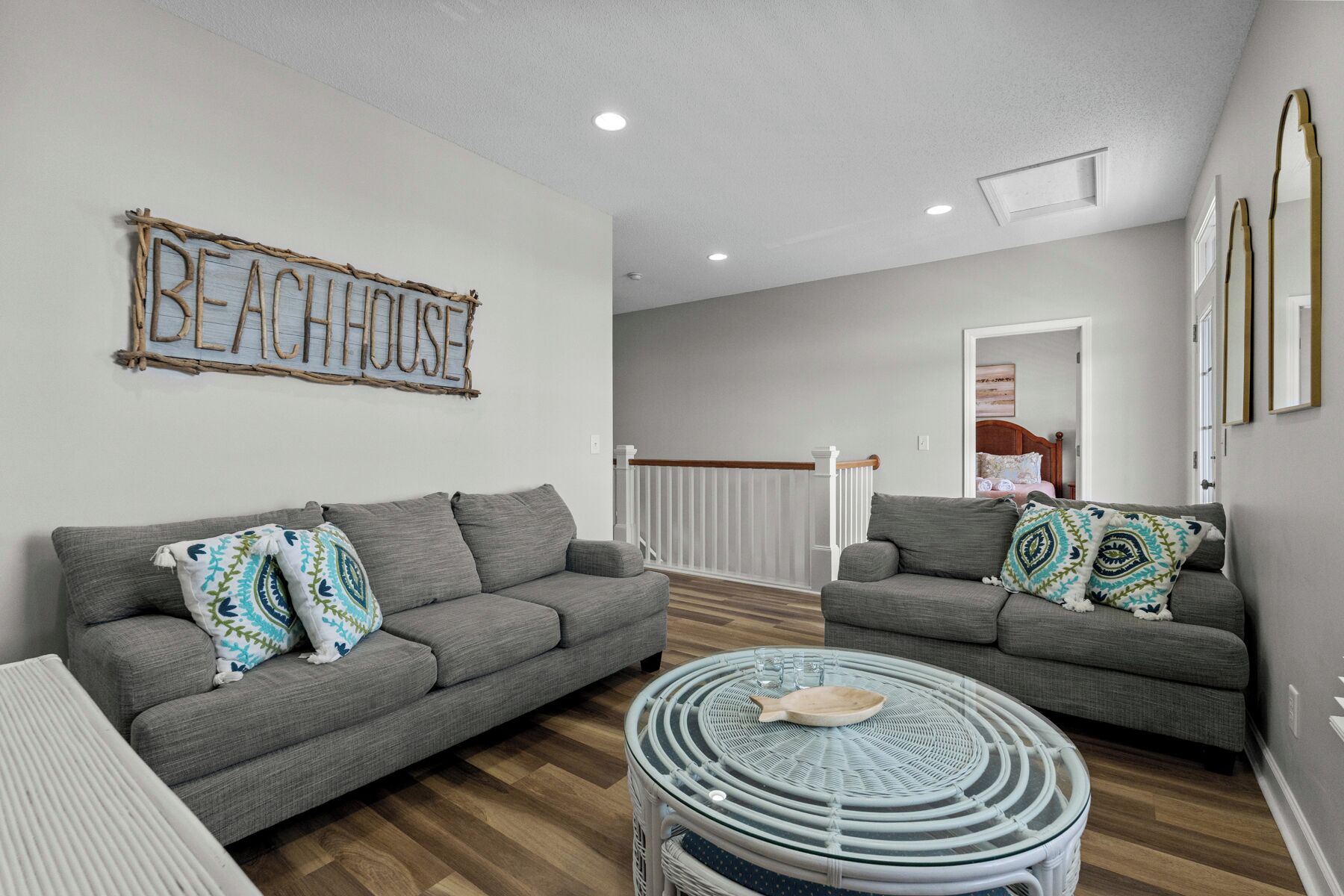 A living room with two grey sofas, a round wicker coffee table, and a 'BEACHHOUSE' sign on the wall.