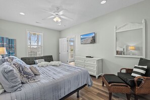 A bedroom with a bed, a chair, a dresser, a ceiling fan, and a beach-themed painting. - Crabby Paddy: Short Walk To Beach, Sunset Views (Garden City Beach)