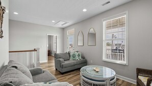 A spacious living room with a grey sofa, a round glass table, and a wicker chair. - Crabby Paddy: Short Walk To Beach, Sunset Views (Garden City Beach)