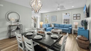 A dining area with a black table, white chairs, and a chandelier. There is a blue sofa and a television in the living area. - Crabby Paddy: Short Walk To Beach, Sunset Views (Garden City Beach)