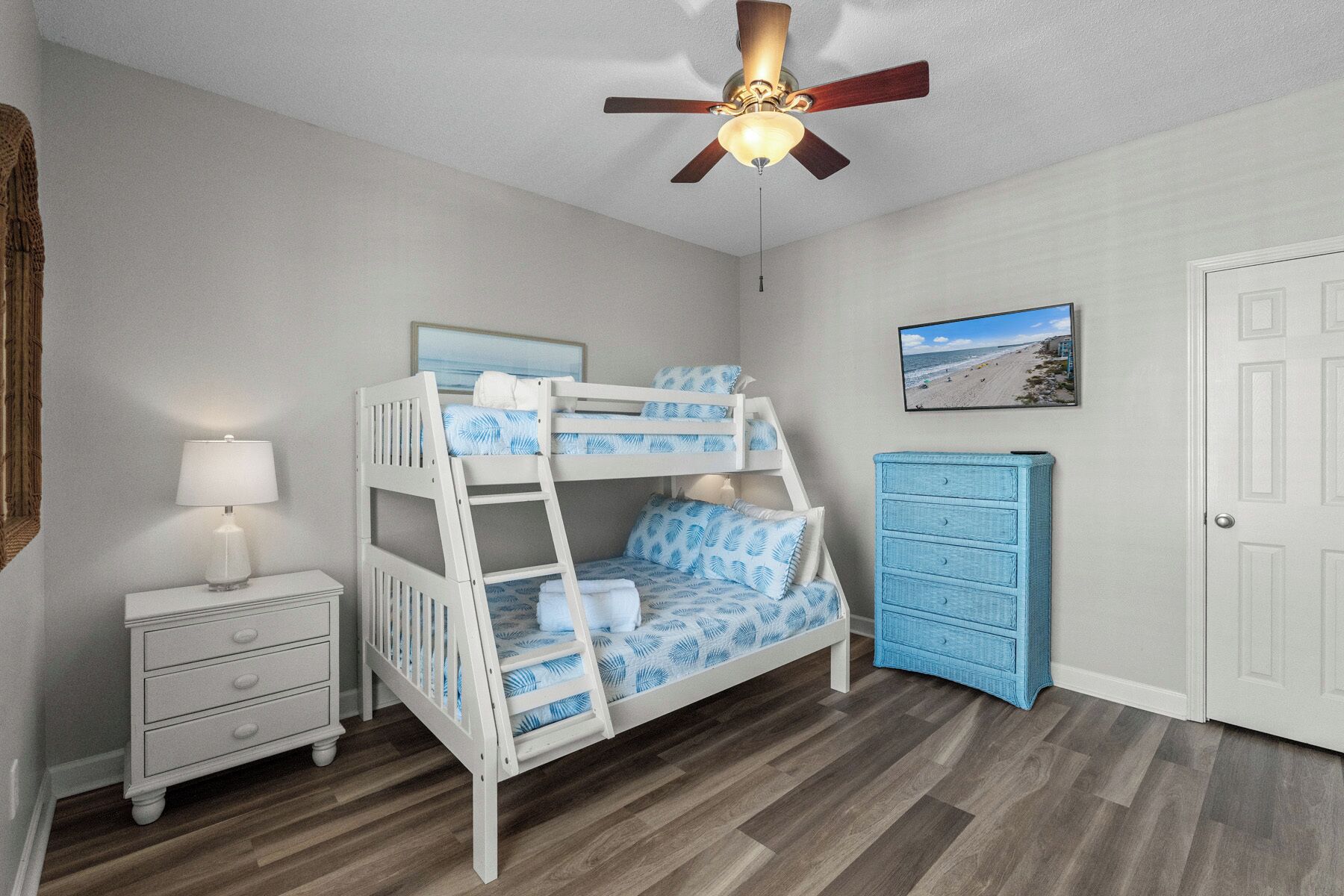 A bunk bed with a ceiling fan, a bedside table with a lamp, a dresser, and a framed picture on the wall.