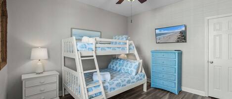 A bunk bed with a ceiling fan, a bedside table with a lamp, a dresser, and a framed picture on the wall.