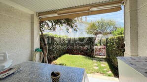 A covered patio area with a table and chairs, a small garden with a hedge, and a view of a building in the background. - House in Golf St. Jordi in La Llosa  (Cambrils)