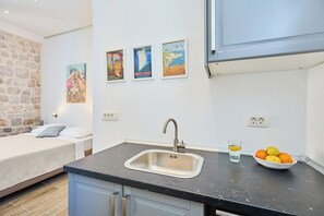 A compact bedroom with a bed, a small kitchenette with a sink and a bowl of fruit, and a stone wall.
