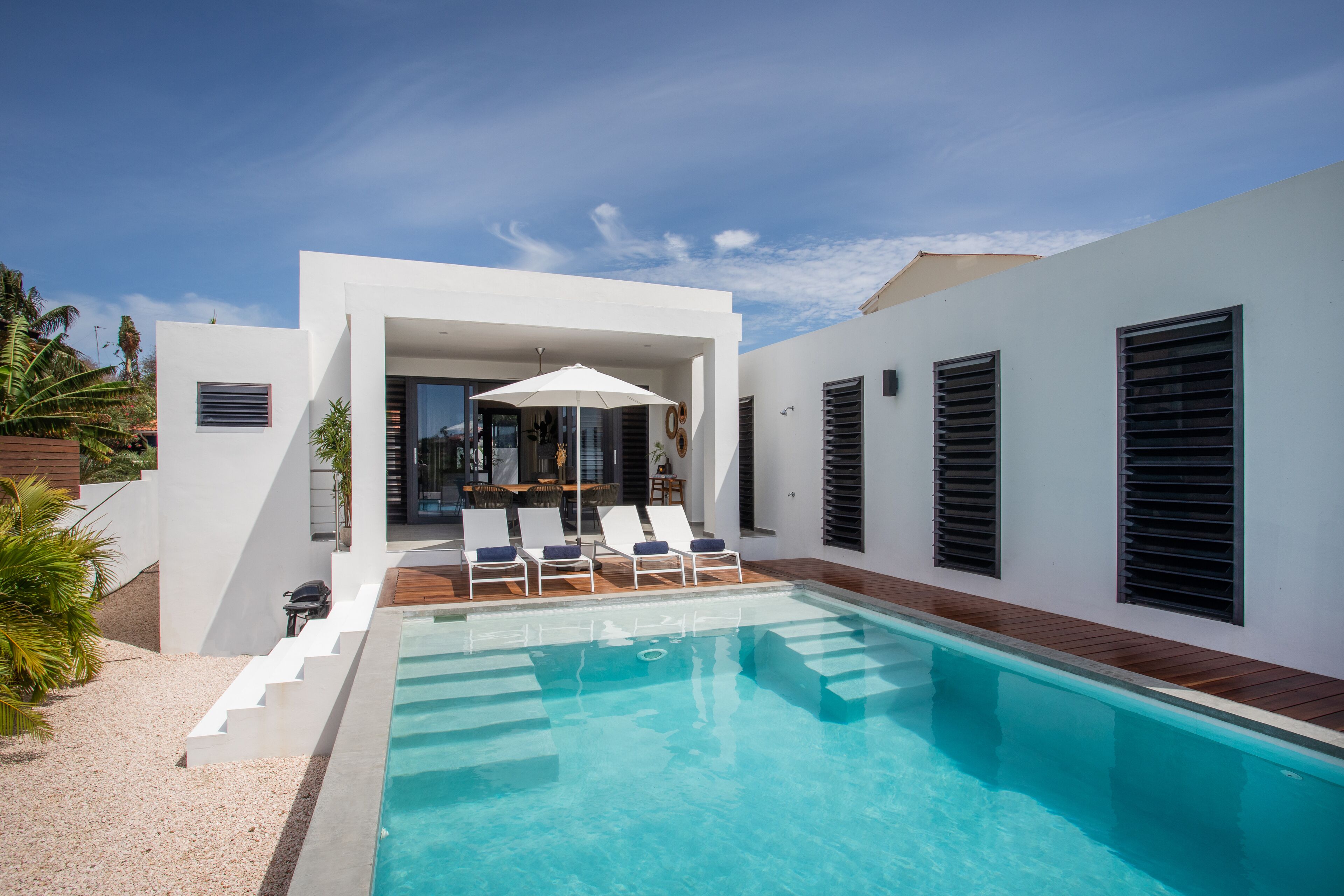 Un espace piscine extérieur moderne, avec une piscine à l’eau d’un bleu limpide, des murs blancs et une terrasse en bois.