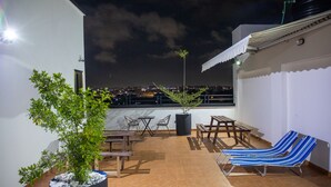 Une terrasse sur le toit offrant une vue sur la ville la nuit, agrémentée d’une plante en pot, de meubles en bois et de chaises longues à rayures.