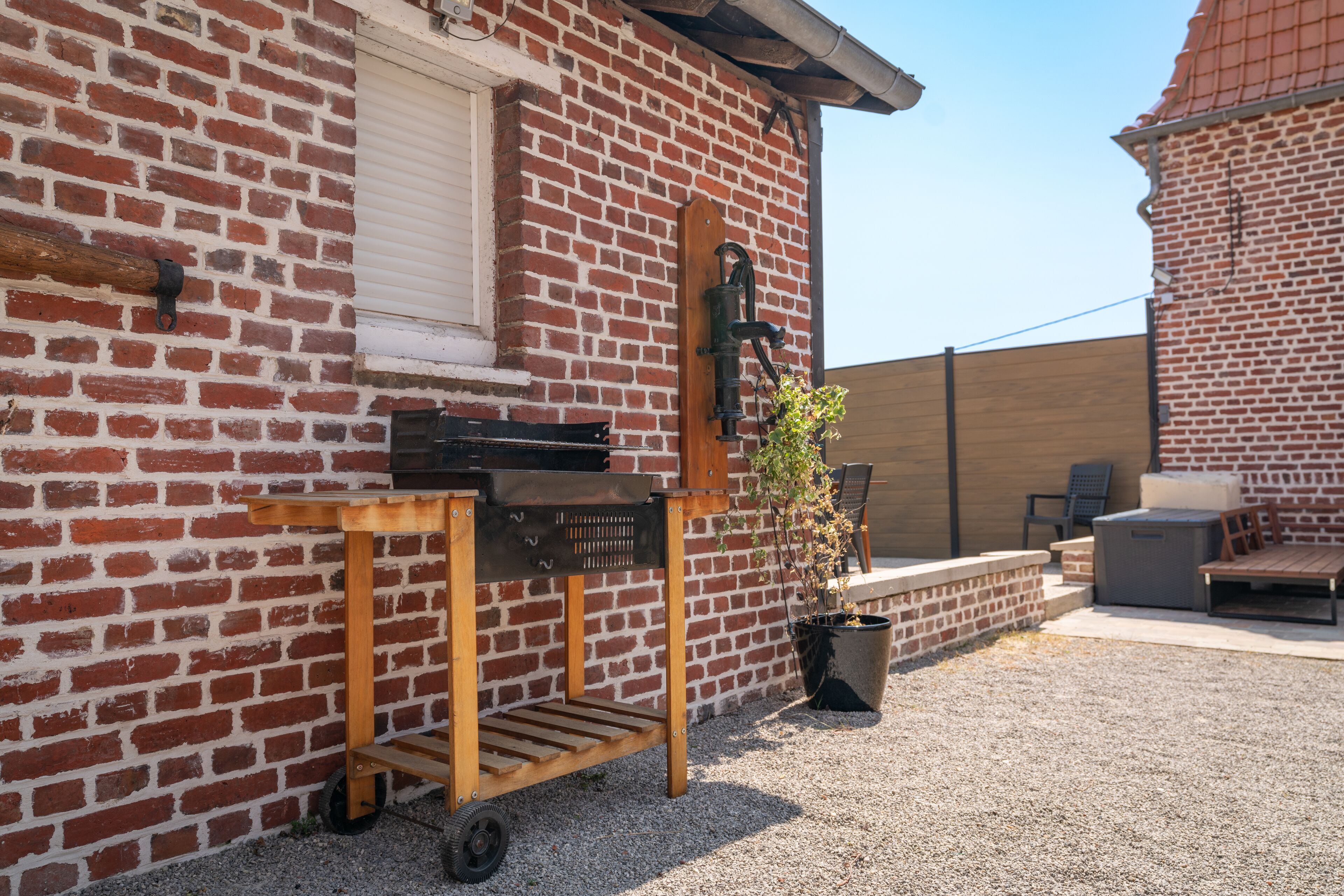 A brick building with a window, a wooden cart with a grill, and a potted plant.