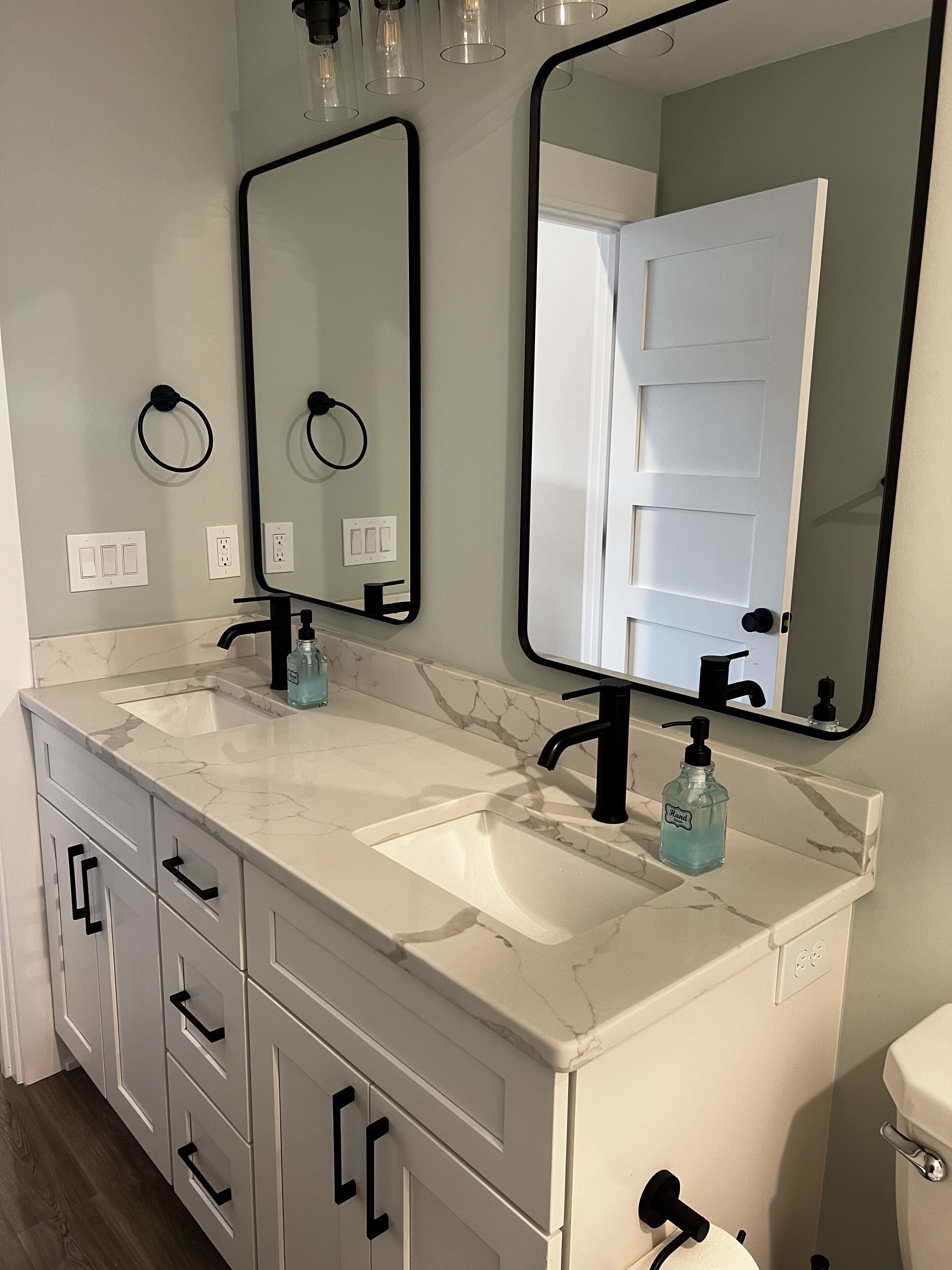 A bathroom with two sinks, a mirror, and a toilet. There are two towel rings on the wall, and a soap dispenser on the sink.