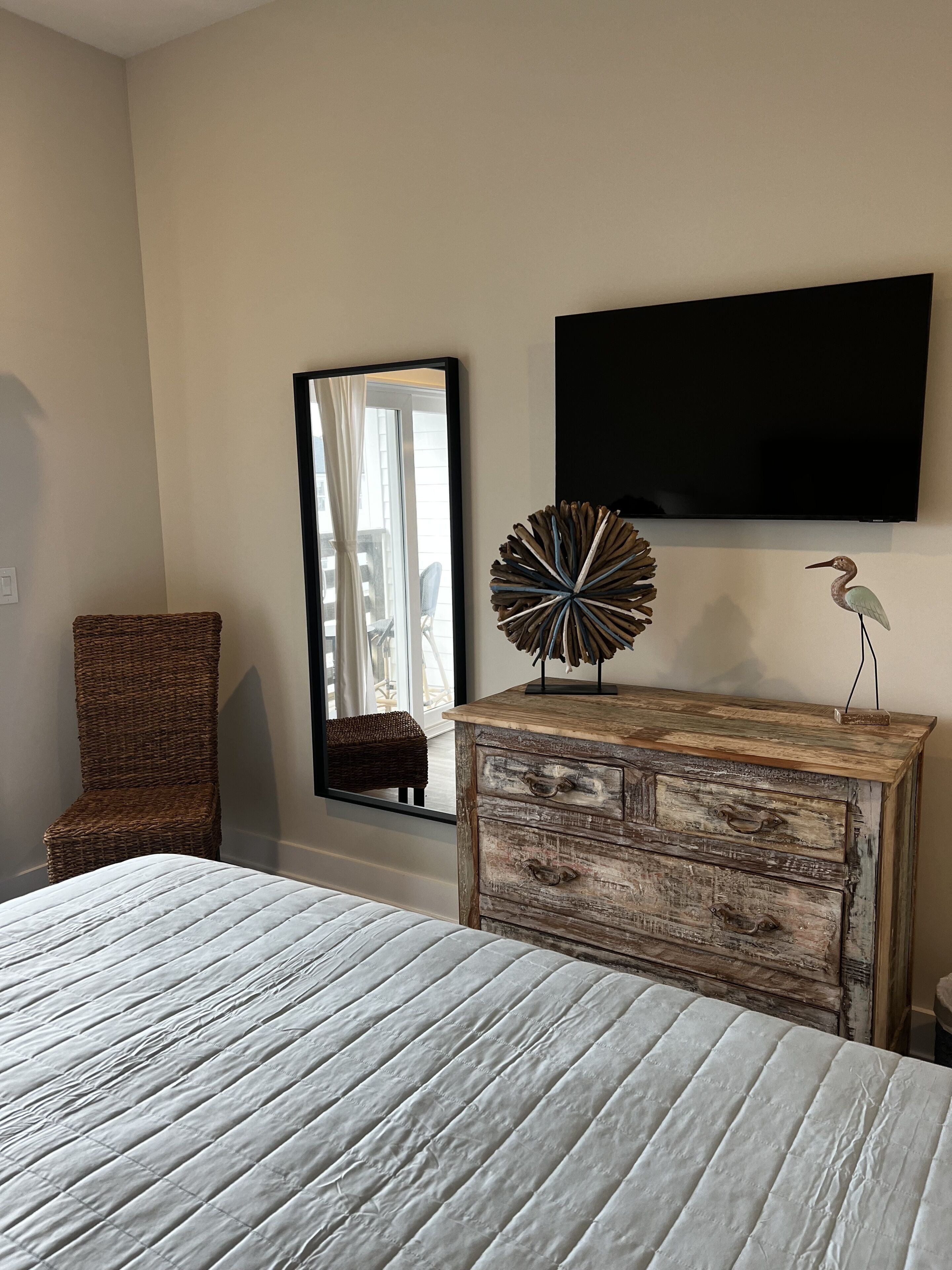 A bedroom with a bed, a wooden dresser, a wicker chair, a mirror, and a flat-screen TV.