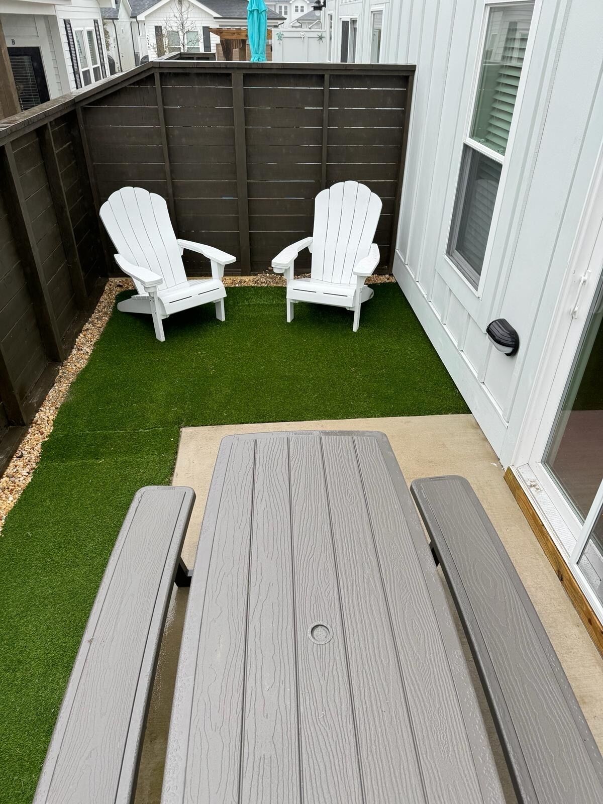 A patio with two Adirondack chairs, a wooden table, and artificial grass.