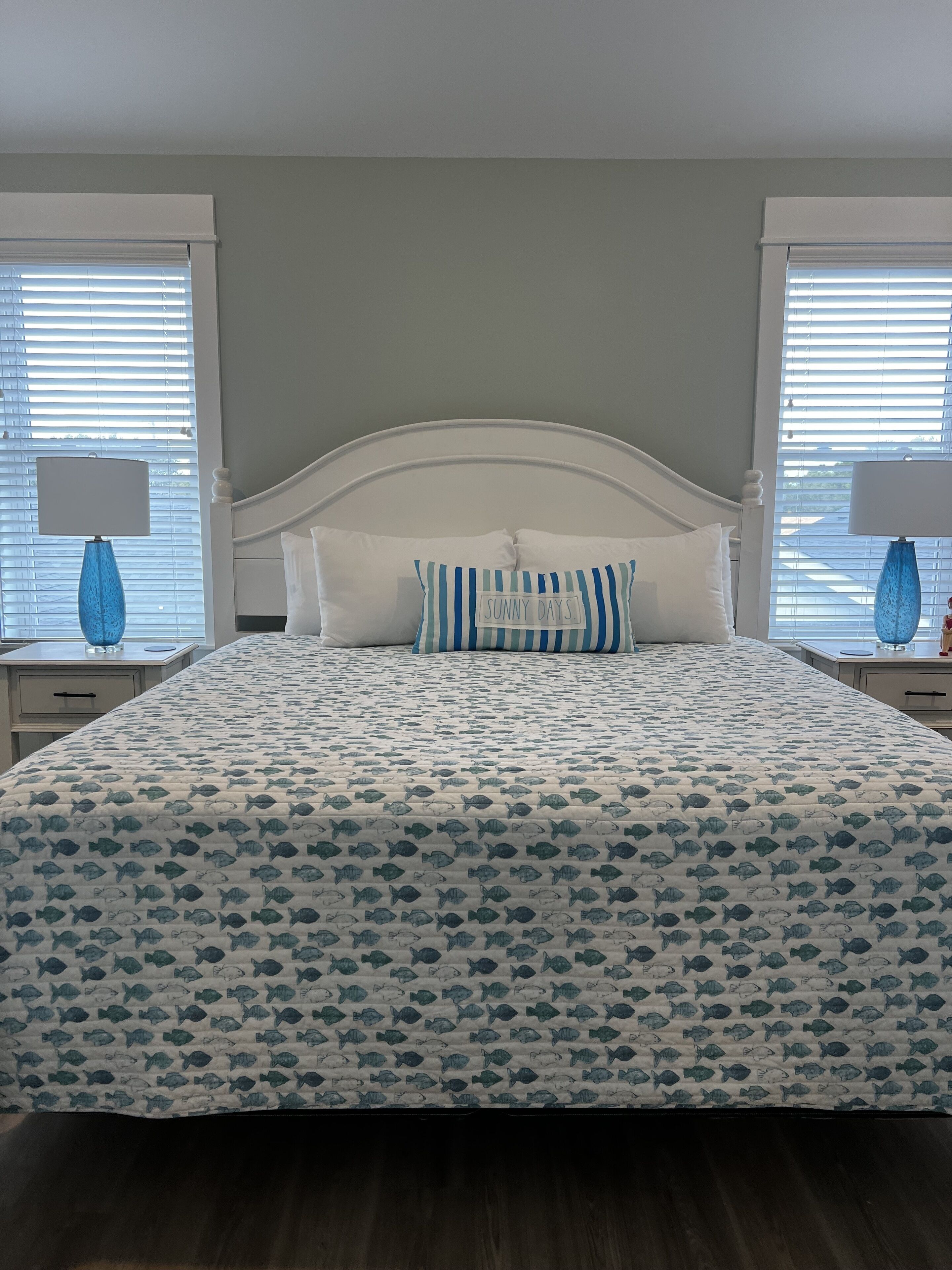 A bed with a white headboard, a blue and white striped pillow, and a bedspread with a fish pattern. There are two bedside tables with lamps and a window with blinds.